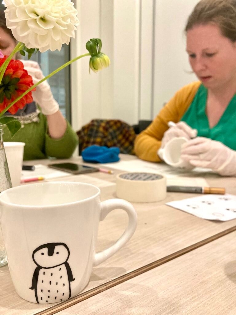 Un mug, décoré avec un petit pinguin, quelqu'un est entrain de décorer un autre mug .