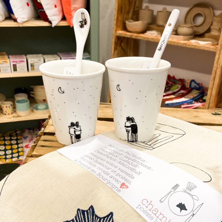 Deux tasses en porcelaine, peint à la main avec une cuillère et une torchon avec une broderie dessus. Derrière il y ades étagères avec pleins de produits, tous fait main